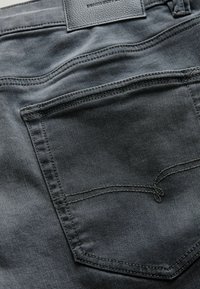 Gray denim fabric showing a close-up of a back pocket with contrasting stitching and a small leather label at the waistband.