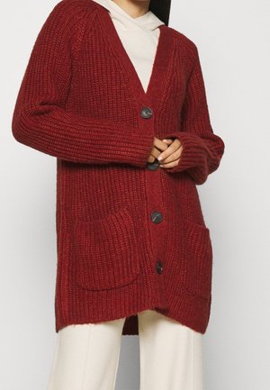 Cardigan tricoté rouge avec un col en V, des poches avant et des boutons noirs. Présente une texture côtelée et une coupe décontractée. Porté par-dessus un sweat à capuche léger.