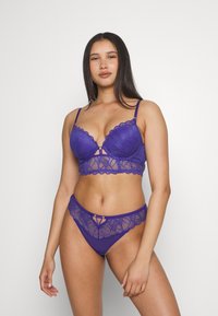 Purple lace bralette with scalloped edges and decorative hardware, paired with matching lace panties featuring a V-shaped accent.