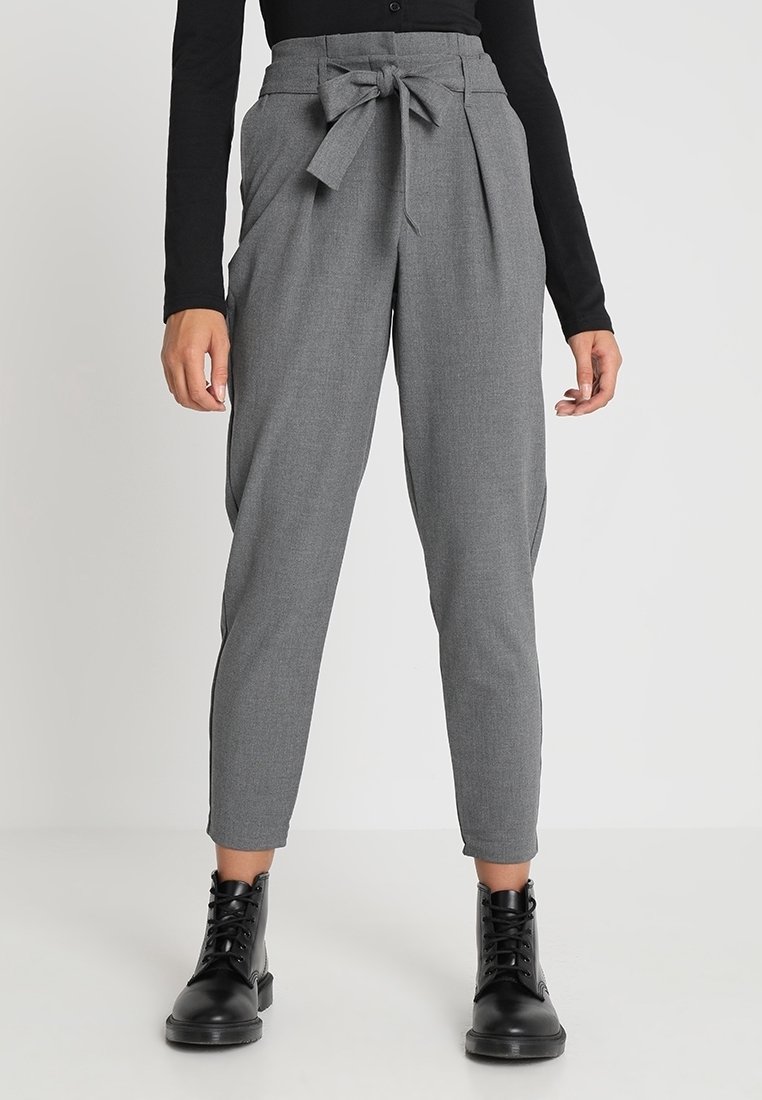 Femme portant un pantalon gris taille haute sur mesure avec un nœud devant, un haut noir à manches longues et des bottines noires à lacets.