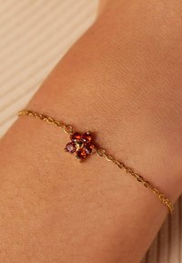 Gouden schakelen armband met een bloemdesign, met oranje en rode edelstenen in het midden, tegen een getextureerde huidachtergrond.