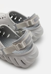 Grå Crocs laget av myk gummimateriale, med ventilasjonshull, teksturert såle og justerbar stoffrem med logodesign.