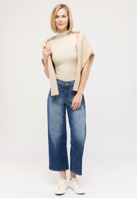 Beige Rollkragenoberteil, leichter, weicher Oversized-Cardigan lässig über die Schultern gelegt, weit geschnittene blaue Jeans, weiße Sneaker. Sanfte Texturen und lässige Passform.
