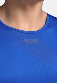 Camiseta deportiva azul hecha de material transpirable, con un cuello redondo y una pequeña textura de malla. Logotipo "2XU" en dorado cerca del cuello.