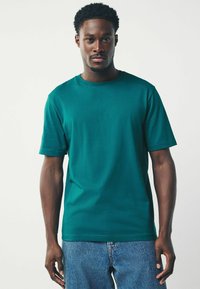 Camiseta de algodón en verde azulado con mangas cortas y escote redondo, combinada con unos jeans de mezclilla azul claro. Textura suave, diseño de corte casual.