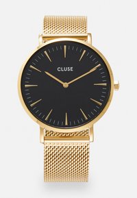 Montre à bande en maillon doré avec un cadran rond noir, des index en or et des aiguilles dorées. Nom de la marque "CLUSE" affiché en blanc sur le cadran.
