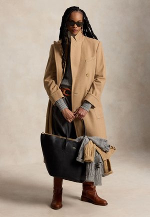 Femme avec de longues tresses portant des lunettes de soleil, un manteau beige, un pull gris, des bottes marron, tenant un sac cabas noir contenant des tricots pliés dans des tons neutres.