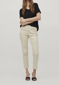 Kvinna iklädd en svart kortärmad V-ringad topp, krämfärgade raka byxor som slutar ovanför anklarna, och svarta öppna klacksandaler som står med armarna korsade.