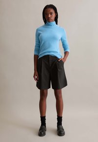Light blue ribbed turtleneck sweater paired with dark gray tailored shorts. Model wears black socks and slip-on shoes, standing against a neutral background.