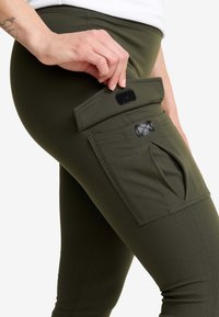 Olivgröna stretchleggins med en sidoficka med kardborrestängning och en liten ytterligare öppningsdetalj, tillverkade av ett mjukt tyg.