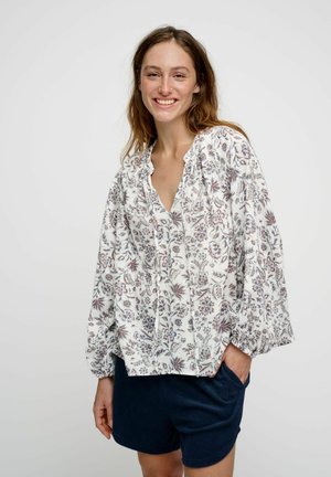 Lachende vrouw die een losse witte bloemenblouse met lange mouwen en marineblauwe shorts draagt, staand met één hand in haar zak.