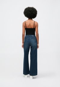 Mulher com cabelo afro, vestindo um top preto, jeans de perna larga azuis e ténis brancos, de pé de costas sobre um fundo simples.
