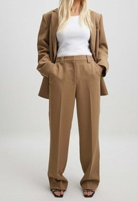 Blazer ajusté beige et pantalon large assorti en tissu texturé. L'ensemble comprend un top blanc cintré et des talons ouverts noirs.