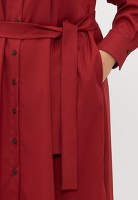 Vestido camiseiro vermelho com cinto, de silhueta reta, apresentando botões pretos na frente e um bolso lateral. Tecido liso e texturado.