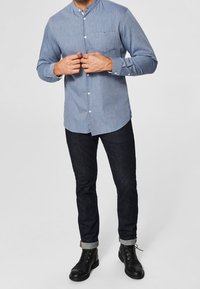 Košile na knoflíky v světle modré barvě s mandarínským límečkem, vybavená náprsní kapsou. V kombinaci s tmavými jeansy a černými šněrovacími botami.