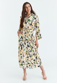 Rochie florală cu mâneci lungi, de culoare crem, cu modele galbene, verzi și negre. Are guler, talie elastică și buzunare laterale. Textură netedă.