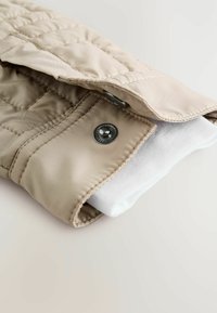 Beige quiltet jakkeærme med en trykknap og hvid ribbet stofmanchet, der strækker sig fra indersiden af ærmet.