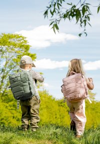 Kaksi lasta, jotka kantavat reppuja ja seisovat nurmikolla aurinkoisessa ulkoilmassa, taustalla puita ja sininen taivas.