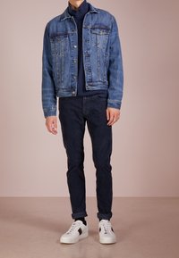 Muž s modrou denimovou bundou přes námořnický svetr na zip, tmavými ohrnutými kalhotami a bílými teniskami se červenými a zelenými pruhy.