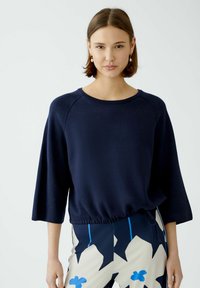 Marineblaues lässiges Sweatshirt mit Rundhalsausschnitt, Dreiviertelärmeln und gerafftem Saum, kombiniert mit gemusterten Hosen, die blaue Akzente aufweisen.