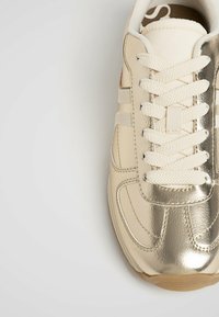 Beige sneaker met een glanzende gouden metalen neuskap, een bovenzijde van getextureerd leer, witte veters en een rubberen zool. Gestikte details langs de zijkanten.