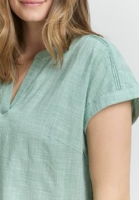 Green checkered top with a v-neckline and short cuffed sleeves. Fabric appears lightweight and textured, featuring a pocket on the left side.