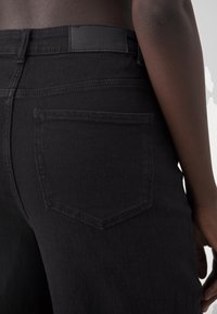 Černé džíny z denimu s jednoduchou zadní kapsou a koženou nášivkou na pase. Tkanina má hladkou strukturu.