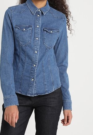 Kvinde iført en tætsiddende blå denim-skjorte med knapper, brystlommer og lange ærmer, kombineret med sorte bukser.