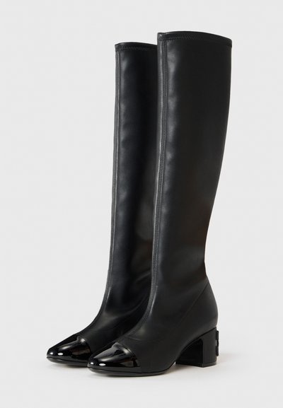 Par de botas negras hasta la rodilla con punteras redondeadas de charol y tacones de bloque sobre un fondo blanco.