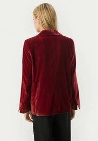 Blazer in velluto bordeaux con colletto, maniche lunghe e taglio dritto. Tessuto testurizzato e dettagli con polsini aperti. Vista posteriore mostrata.