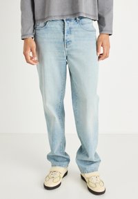 Απαλά μπλε τζιν παντελόνια με ίσια γραμμή και διακριτική φθορά, με πέντε τσέπες και κλασικό κούμπωμα με κουμπί και φερμουάρ. Φοριούνται με μπεζ αθλητικά παπούτσια.