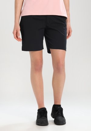 Persoon draagt zwarte knielange shorts, lichtroze shirt en zwarte sportschoenen, staand op een effen achtergrond.