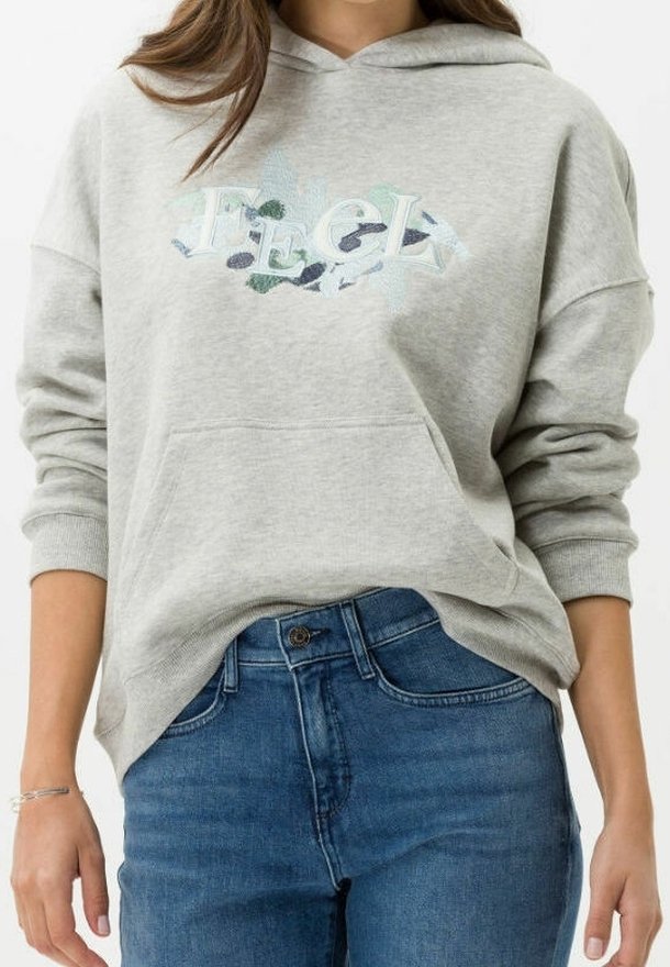 Femme portant un sweat à capuche gris clair avec le texte "FEEL" et un motif abstrait, associée à un jean en denim bleu.