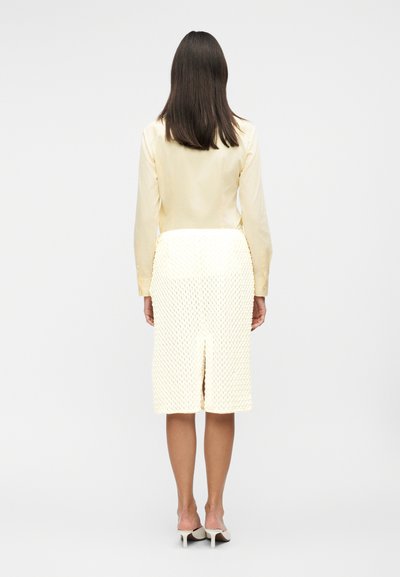 Γυναίκα με μακριά σκουρόχρωμα μαλλιά στέκεται πλάτη, φορώντας ανοιχτό κίτρινο πουκάμισο, υφασμάτινη λευκή φούστα μήκους μέχρι το γόνατο και λευκά ψηλοτάκουνα σε λευκό φόντο.