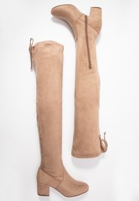 Botas nude de ante por encima de la rodilla. Presentan un tacón cuadrado y cierre de cremallera lateral. Cinta ajustable en la parte superior para un ajuste ceñido.