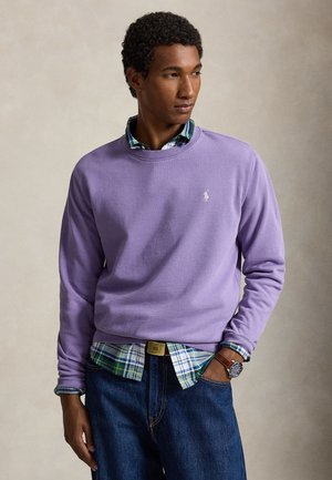 Mann trägt einen lavendelfarbenen Pullover über einem grün-blau karierten Hemd, blaue Jeans und eine braune Uhr, steht mit einer Hand in der Tasche.