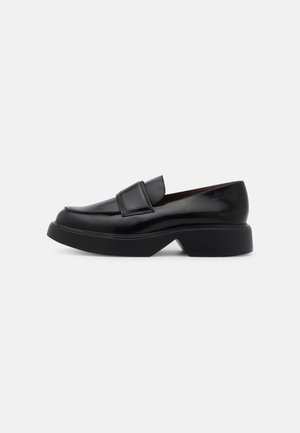 Mocassins en cuir noir avec une semelle plateforme épaisse, présentant un dessus cousu et un détail de sangle à l'avant. Texture lisse, design minimaliste.