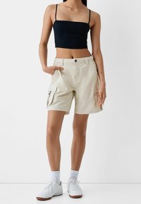 Beige cargo šortky s bočními kapsami, v kombinaci s přiléhavým černým crop topem a bílými teniskami. Materiál působí lehkým a neformálním dojmem.