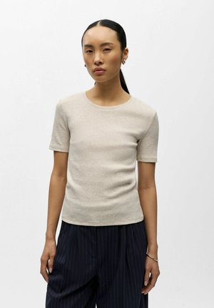 Kvinne med langt svart hår samlet bak, iført en beige kortermet topp og marineblå vide bukser med tynne striper, mot en ensfarget bakgrunn.