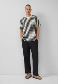 Chemise à rayures à manches courtes avec des rayures horizontales noires et crème, associée à un pantalon noir ample et des sandales marron avec boucle.
