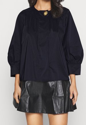 Vrouw draagt een zwarte blouse met lange mouwen en een gouden knoop bij de hals, gecombineerd met een korte zwarte leren rok.
