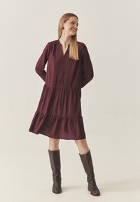 Pika-sleeve, põlvikõrgune burgundi värvi kleit geomeetrilise mustriga. Omab V-kaelust, kihilist seelikuosa ja kerget kangast. Kooritud pruuni põlvini ulatuva saapaga.