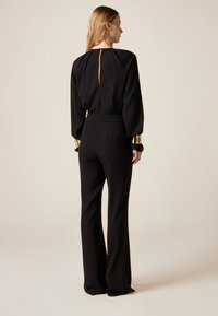 Musta haalari, jossa on pitkät hihat, korkea kaulus ja takana avonainen keyhole-leikkaus. Hihansuissa kultaiset koristenapit.