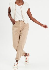 Beige högmidjade byxor, vit t-shirt och en krämfärgad texturerad väst med knappar, i kombination med sneakers med orange detaljer.