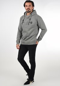 Grauer Kapuzenpullover mit Kordeln, schwarzem Patch-Logo, kombiniert mit schwarzen Jeans und schwarzen Sneakers mit goldenen Akzenten.