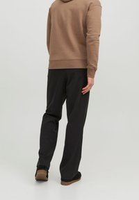 Sudadera de tela marrón claro con puños y dobladillo acanalados, combinada con pantalones negros. El atuendo incluye calzado negro minimalista con una suela texturizada.