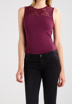 Kvinde iført ærmeløs bordeaux top med blonde detaljer over brystet og sorte højtaljede jeans, stående foran en ensfarvet baggrund.