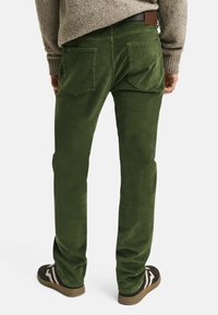 Persona con pantalones de pana verdes con bolsillos traseros, suéter beige y zapatillas negras con rayas blancas, de pie de espaldas.