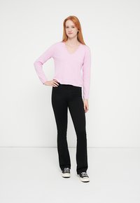 Ροζ ανοιχτού χρώματος, μακρυμάνικη μπλούζα με V-λαιμόκοψη, συνδυασμένη με μαύρο καμπάνα παντελόνι και μαύρα αθλητικά παπούτσια, παρουσιασμένη πάνω σε λευκό φόντο.