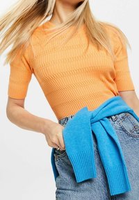 Oranžový žebrovaný pletený top s krátkými rukávy, doplněný modrým pleteným svetrem uvázaným kolem pasu. Viditelné denimové šortky.
